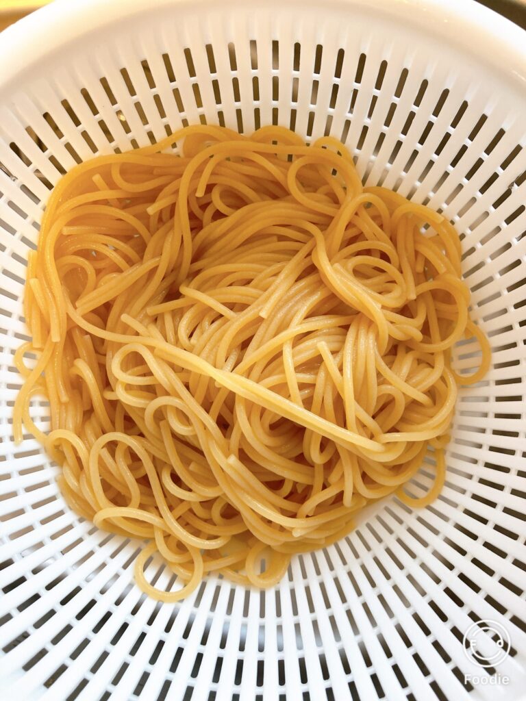 茹で上がったゼンブヌードルのスパゲッティ麺をザルにあげた様子。ほんのり黄色みがかった色合いと、つやのある表面が特徴。