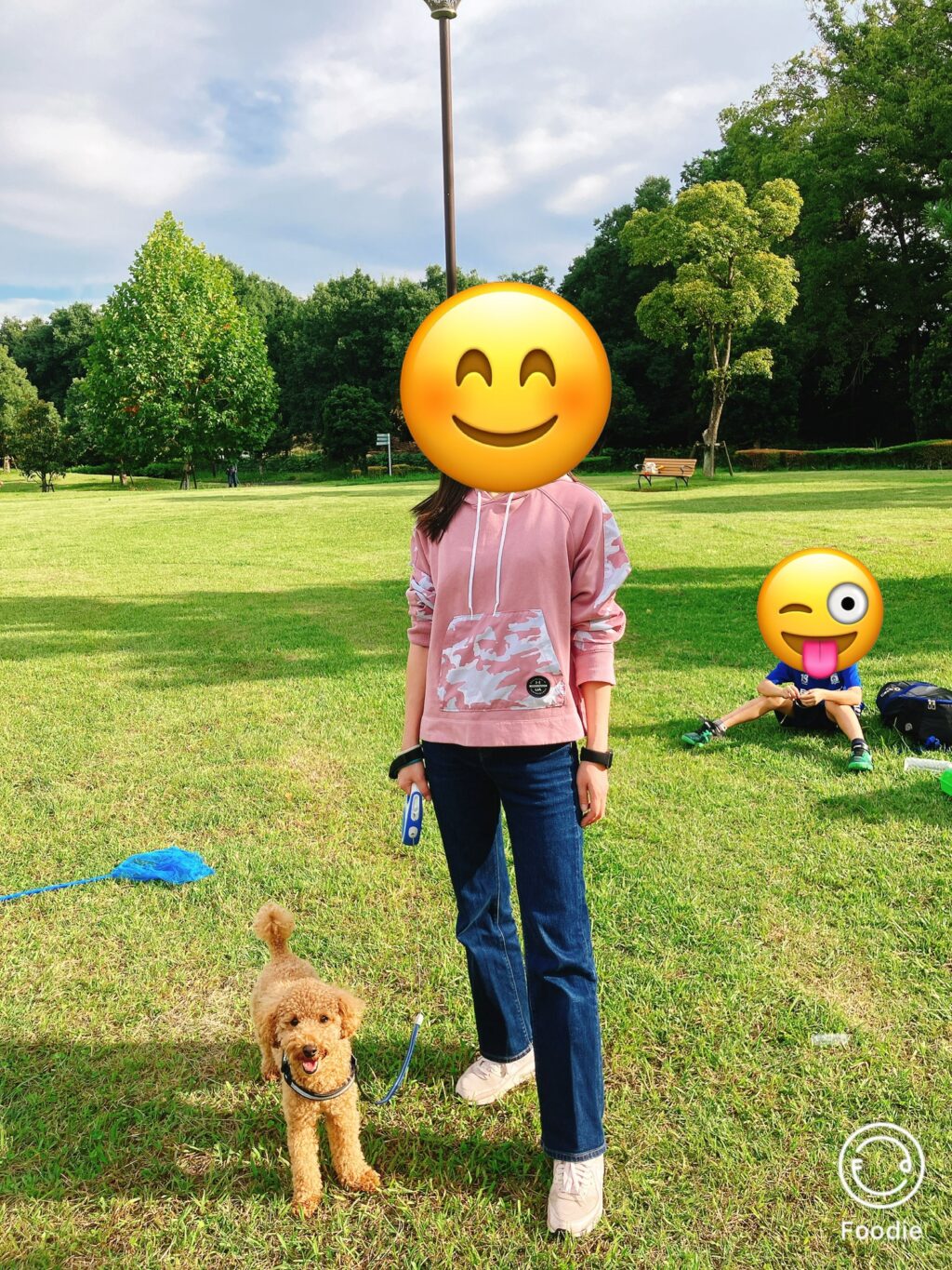 秋の公園でピンクのパーカーとジーンズを着た女性が犬と一緒に立っている様子。晴れ間がのぞき、緑の芝生が広がる中でリラックスした雰囲気。